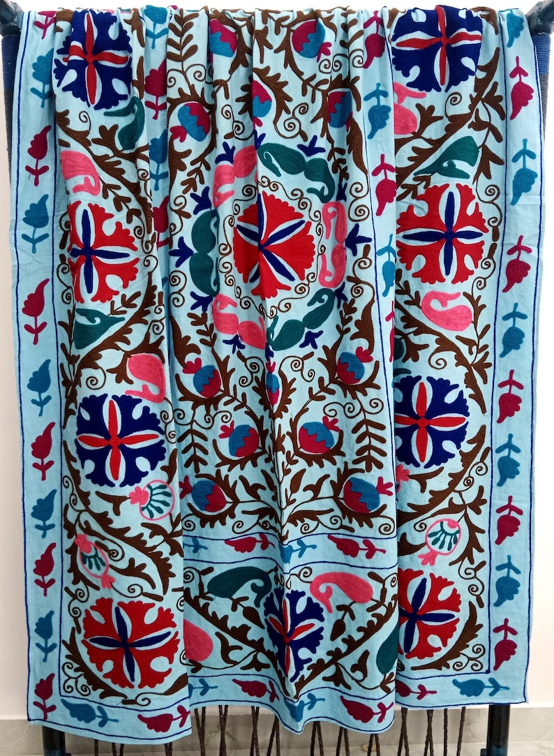 May include: A light blue textile with intricate embroidery. The design features red, blue, pink, and green floral and paisley motifs on a light blue background, with brown vines and a blue border. The textile is likely a decorative throw or blanket.