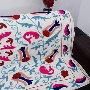 May include: White textile with embroidered floral patterns in shades of pink, blue, and red. The design features stylized flowers and leaves, creating a vibrant and decorative look. The textile is draped over a dark surface.
