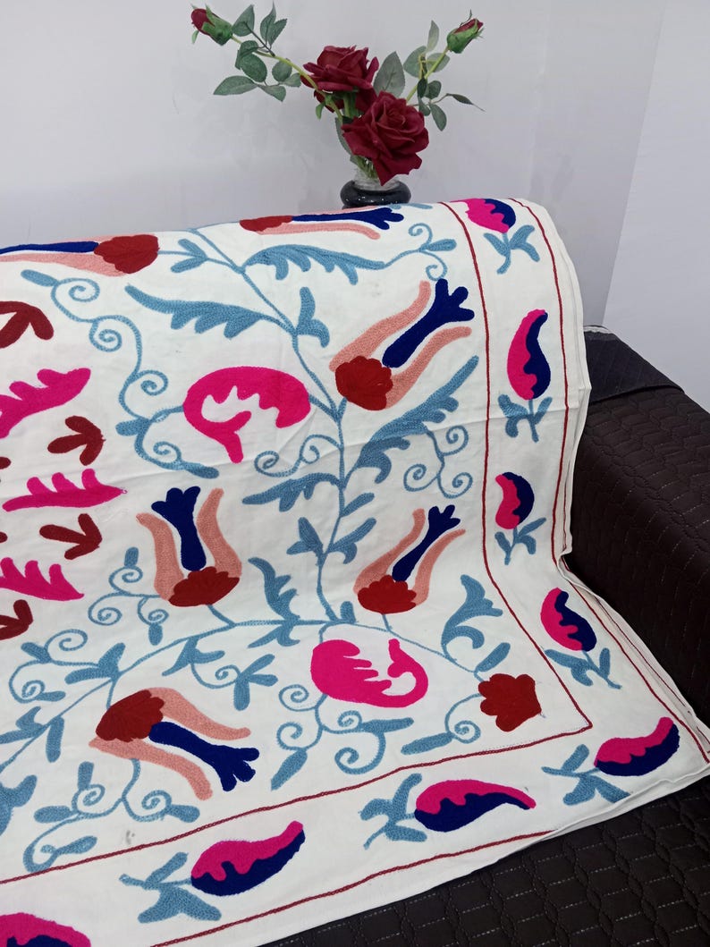 May include: White textile with embroidered floral patterns in shades of pink, blue, and red. The design features stylized flowers and leaves, creating a vibrant and decorative look. The textile is draped over a dark surface.