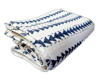 May include: Folded white quilt with a repeating pattern of dark blue triangles. The quilt is stitched with dark thread, and the edges are neatly finished. The fabric appears to have a textured surface.