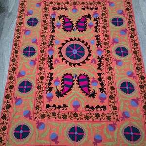 May include: A rectangular, coral-colored rug with intricate embroidery. The design features floral and paisley motifs in shades of pink, blue, green, and brown. The rug has a detailed border and central medallion.