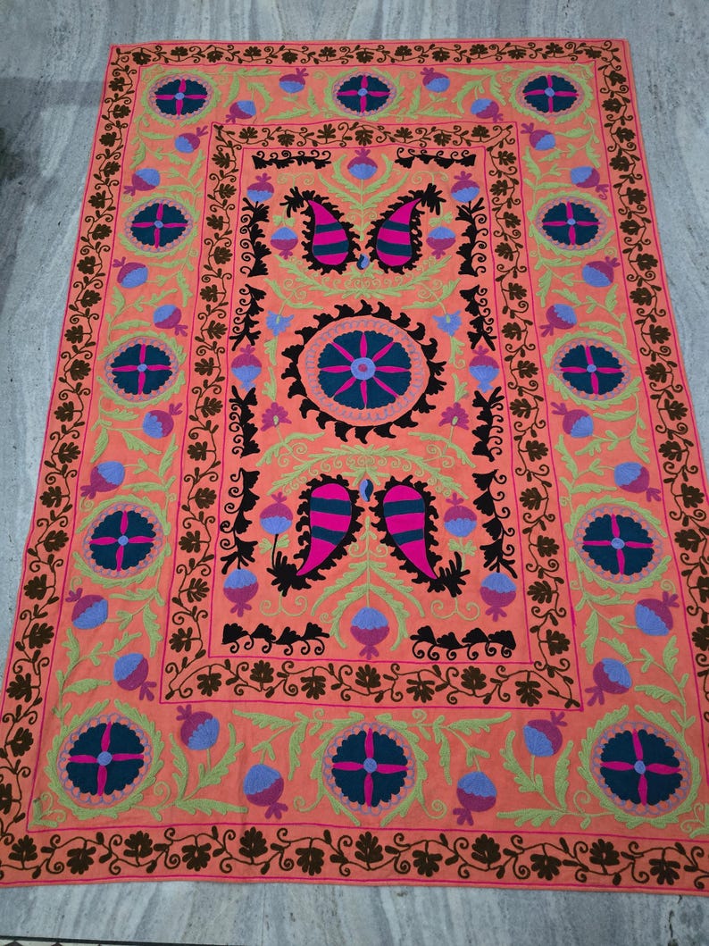 May include: A rectangular, coral-colored rug with intricate embroidery. The design features floral and paisley motifs in shades of pink, blue, green, and brown. The rug has a detailed border and central medallion.
