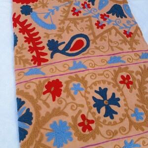 May include: Peach-colored textile with intricate embroidery. The design features floral and paisley motifs in shades of blue, red, and gold. A border with a pink stripe runs along the edge. The textile is likely a decorative item, such as a tablecloth or wall hanging.