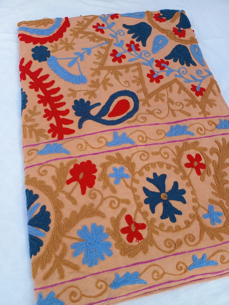 May include: Peach-colored textile with intricate embroidery. The design features floral and paisley motifs in shades of blue, red, and gold. A border with a pink stripe runs along the edge. The textile is likely a decorative item, such as a tablecloth or wall hanging.