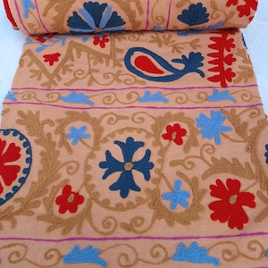 May include: A rolled-up peach-colored textile with intricate embroidery. The design features floral and paisley motifs in red, blue, and gold thread. The embroidery is detailed and covers the fabric with a repeating pattern.