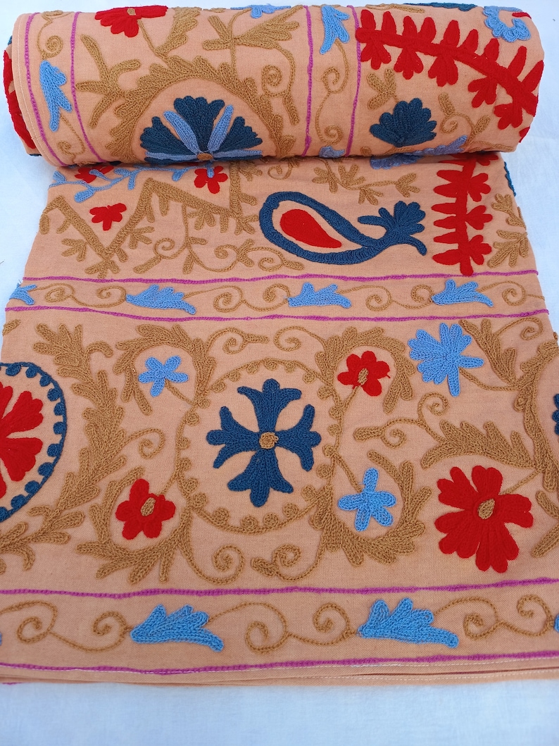 May include: A rolled-up peach-colored textile with intricate embroidery. The design features floral and paisley motifs in red, blue, and gold thread. The embroidery is detailed and covers the fabric with a repeating pattern.