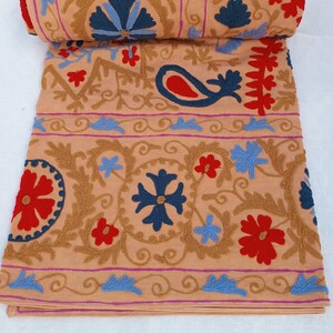 May include: A rolled-up, tan textile with intricate embroidery. The design features blue, red, and gold floral and paisley patterns. The edges are trimmed with a pink border. The textile is folded and rolled, showcasing the detailed craftsmanship.