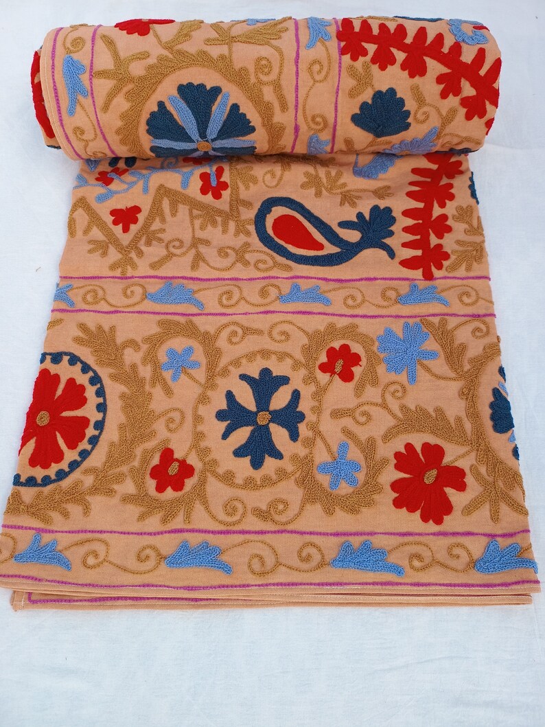May include: A rolled-up, tan textile with intricate embroidery. The design features blue, red, and gold floral and paisley patterns. The edges are trimmed with a pink border. The textile is folded and rolled, showcasing the detailed craftsmanship.