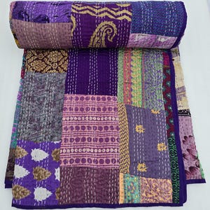 May include: A patchwork quilt with a rolled top, featuring a variety of purple, lavender, and gold fabrics. The quilt is made of rectangular patches stitched together with white thread. The edges are finished with a dark purple border.