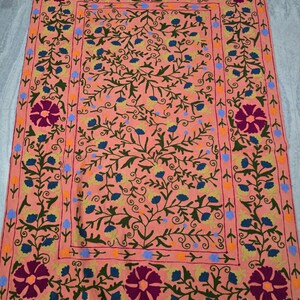 May include: Peach-colored embroidered textile with a floral design. The textile features a central field with a repeating pattern of blue and yellow flowers and green vines. The border has large, dark purple flowers and a repeating pattern.