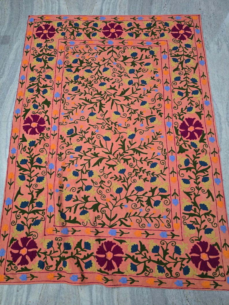 May include: Peach-colored embroidered textile with a floral design. The textile features a central field with a repeating pattern of blue and yellow flowers and green vines. The border has large, dark purple flowers and a repeating pattern.