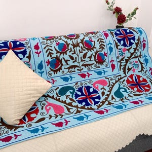 May include: A light blue sofa cover with an intricate embroidered floral design in red, pink, and blue. A cream-colored pillow with a quilted pattern rests on the sofa. The sofa is covered with a white quilted fabric.