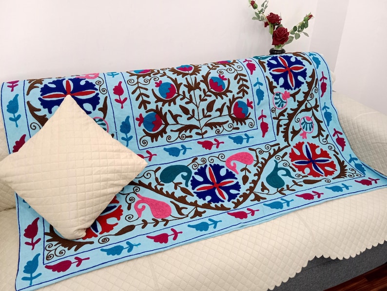 May include: A light blue sofa cover with an intricate embroidered floral design in red, pink, and blue. A cream-colored pillow with a quilted pattern rests on the sofa. The sofa is covered with a white quilted fabric.