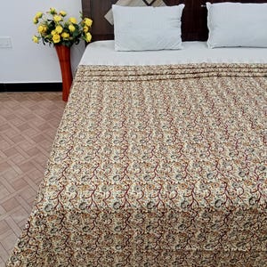May include: A bed with a floral patterned quilt in cream, brown, and dark red. Two white pillows rest against a dark wood headboard. A red vase with yellow flowers sits to the left of the bed.