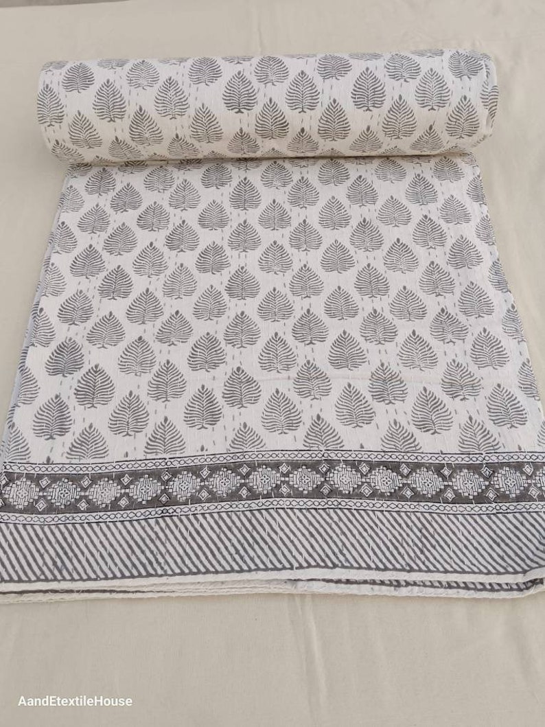 May include: A white cotton fabric with a gray leaf pattern and a gray border. The fabric is rolled up and has a white background.