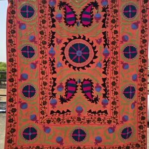 May include: A vibrant, orange textile with intricate embroidery. The design features floral and paisley motifs in shades of pink, blue, green, and black. The textile is rectangular and appears to be a decorative piece, possibly a wall hanging or a throw.
