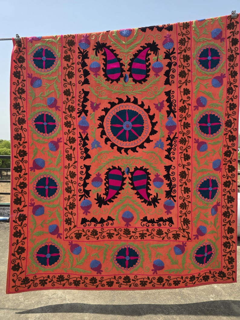 May include: A vibrant, orange textile with intricate embroidery. The design features floral and paisley motifs in shades of pink, blue, green, and black. The textile is rectangular and appears to be a decorative piece, possibly a wall hanging or a throw.