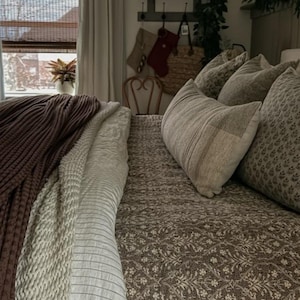 May include: A bed featuring a brown patterned quilt, a white and grey striped blanket, and a dark brown chunky knit throw. Several pillows are arranged on the bed, including some with a small floral print and others with a neutral textured fabric.