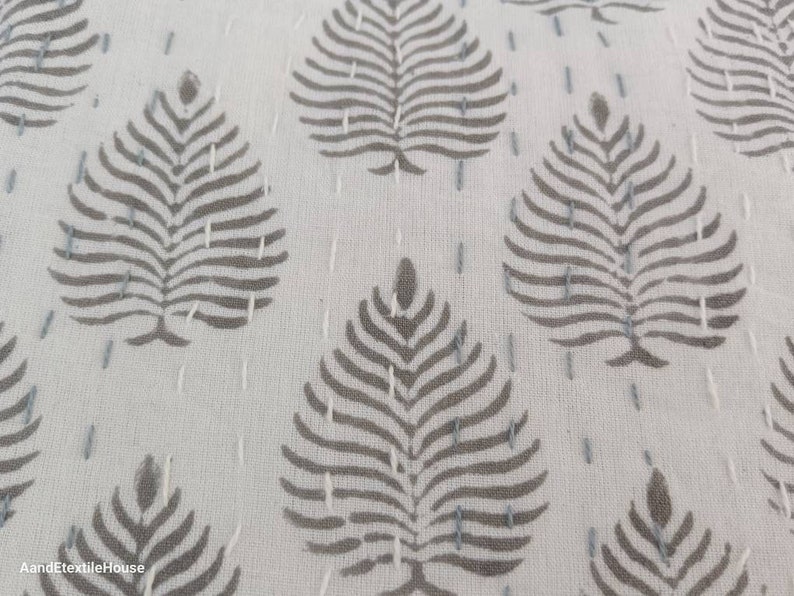May include: Close-up of a white fabric with a repeating pattern of gray leaf designs. The fabric is stitched with small, white stitches.