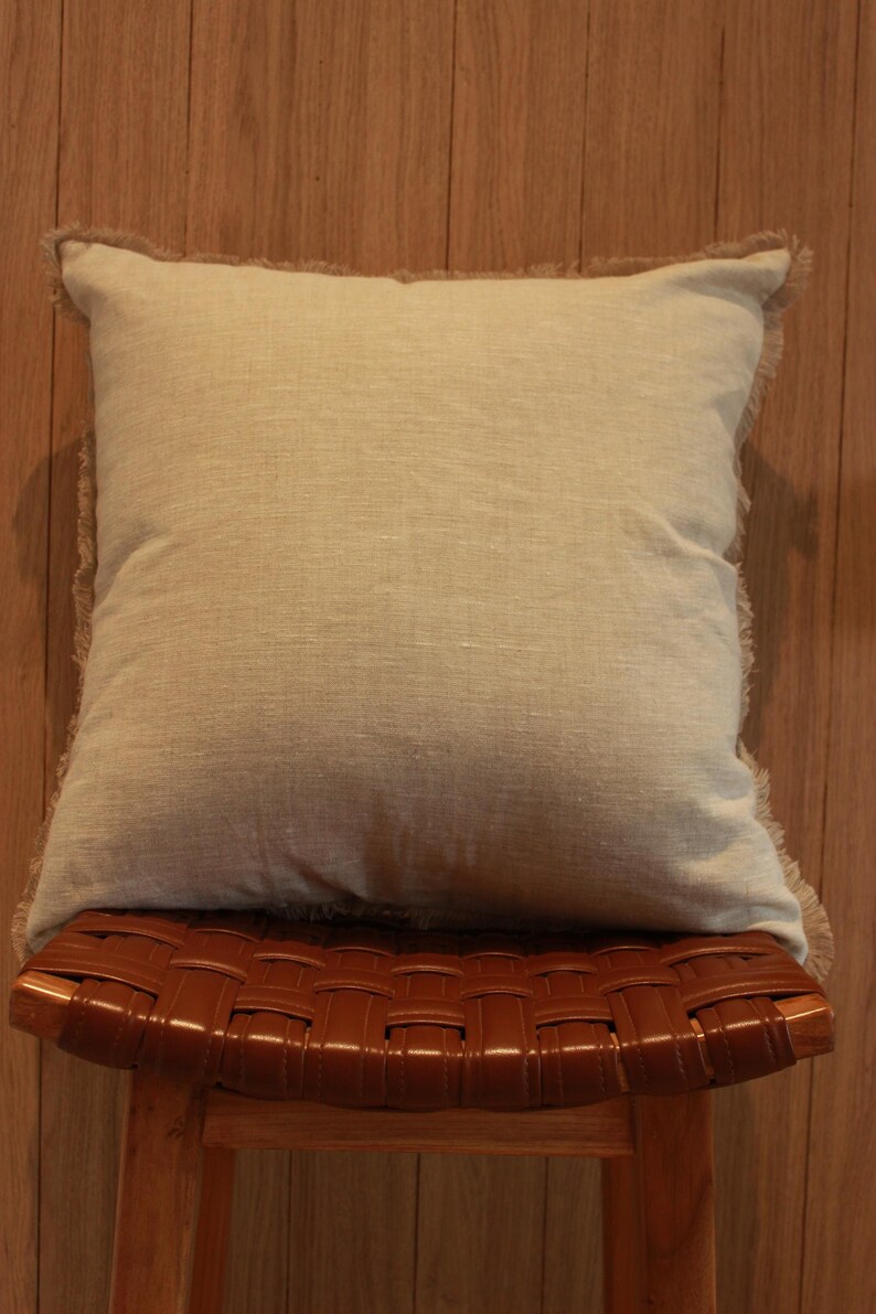 May include: A light beige throw pillow with a frayed edge sits on a brown leather woven stool. The stool has a wooden frame and legs.