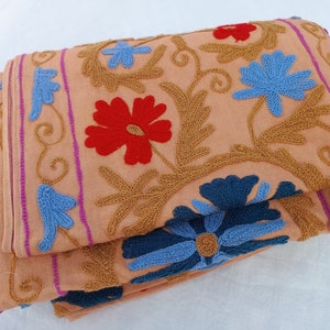 May include: Folded peach-colored fabric with intricate embroidery. The design features red, blue, and gold floral patterns. The edges are trimmed with pink and purple stripes. The fabric is folded over, showcasing the detailed craftsmanship.