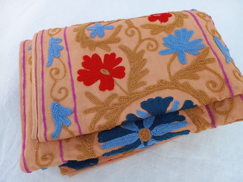 May include: Folded peach-colored fabric with intricate embroidery. The design features red, blue, and gold floral patterns. The edges are trimmed with pink and purple stripes. The fabric is folded over, showcasing the detailed craftsmanship.