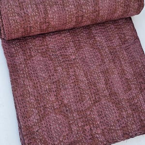 May include: A rolled-up, burgundy-colored quilt with a floral pattern and white stitching. The quilt is partially unrolled, revealing the detailed stitching and design. The fabric has a slightly wrinkled texture.