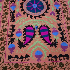 May include: A peach-colored textile with intricate embroidery. The design features a central circular motif with blue and pink accents, surrounded by black, green, and purple floral patterns. The borders are adorned with repeating floral and leaf patterns.