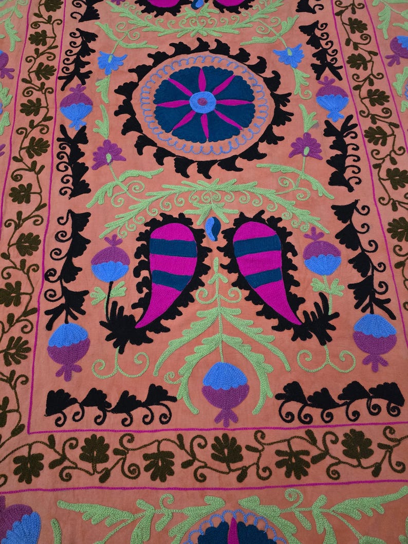 May include: A peach-colored textile with intricate embroidery. The design features a central circular motif with blue and pink accents, surrounded by black, green, and purple floral patterns. The borders are adorned with repeating floral and leaf patterns.
