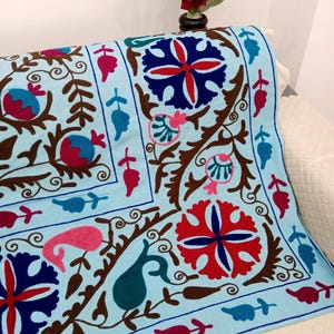 May include: A light blue decorative textile with an embroidered floral pattern. The design features red, pink, and blue flowers, along with brown stems and leaves. The textile is draped over a white surface.