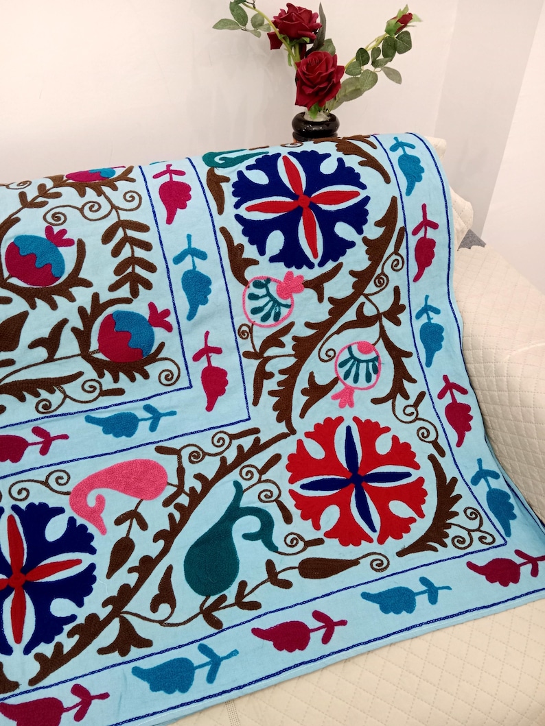 May include: A light blue decorative textile with an embroidered floral pattern. The design features red, pink, and blue flowers, along with brown stems and leaves. The textile is draped over a white surface.