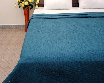 Könnte beinhalten: Eine tealfarbene, gesteppte Tagesdecke bedeckt ein Bett mit weißen Kissen und einem dunklen Holz-Kopfteil. Links vom Bett steht eine hohe orangefarbene Vase mit gelben Blumen. Die Tagesdecke hat eine strukturierte Oberfläche.