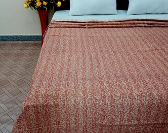 May include: A beige and red patterned kantha quilt is spread out on a bed with two white pillows. The quilt has a floral pattern and is made of cotton.