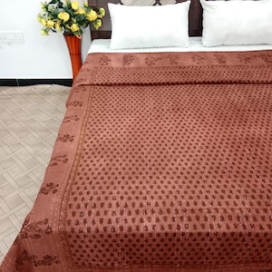 May include: A rust-colored bedspread with a floral pattern covers a bed. The bedspread has a detailed border with floral designs. Two white pillows rest on the bed, and a vase of yellow flowers sits nearby.
