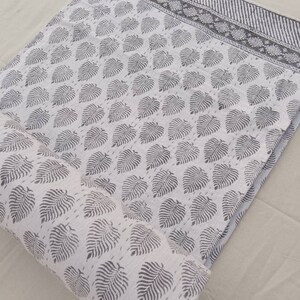 May include: White cotton fabric with a gray leaf print. The fabric is rolled up and has a gray border with a striped pattern.