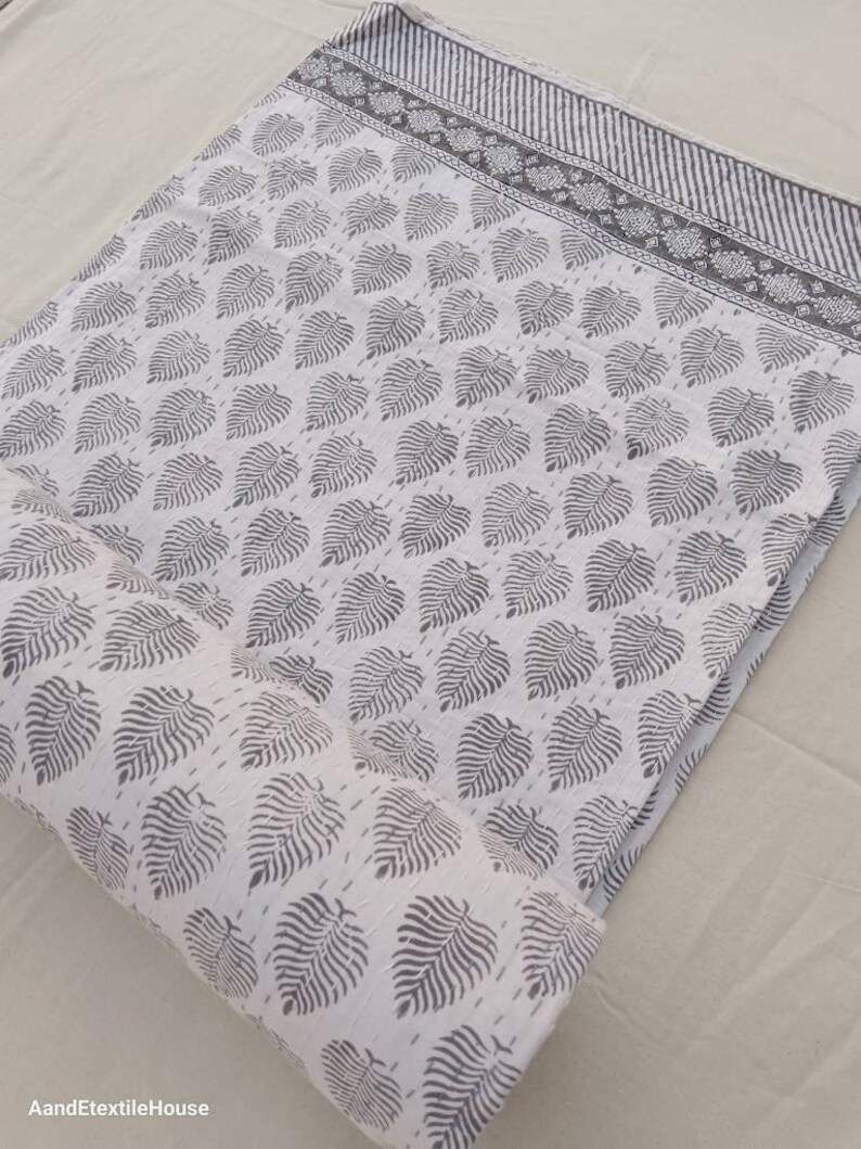 May include: White cotton fabric with a gray leaf print. The fabric is rolled up and has a gray border with a striped pattern.
