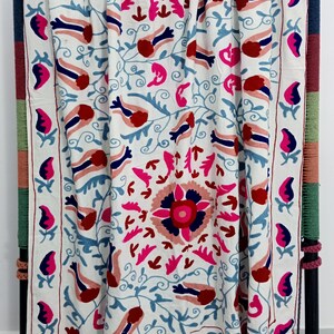 May include: White textile with intricate embroidery featuring a floral and bird design in shades of red, pink, and blue. The border has a repeating pattern of stylized flowers. The textile is draped over a dark metal frame.