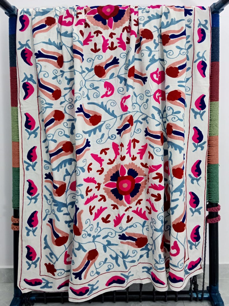 May include: White textile with intricate embroidery featuring a floral and bird design in shades of red, pink, and blue. The border has a repeating pattern of stylized flowers. The textile is draped over a dark metal frame.