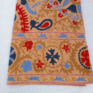 May include: A tan textile with intricate embroidery. The design features red, blue, and gold floral and paisley patterns. The edges are bordered with a repeating design in blue and gold. The textile is folded and lies on a white surface.