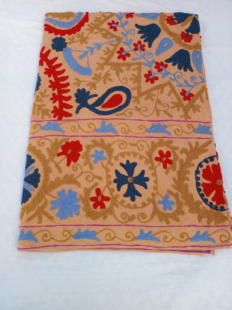 May include: A tan textile with intricate embroidery. The design features red, blue, and gold floral and paisley patterns. The edges are bordered with a repeating design in blue and gold. The textile is folded and lies on a white surface.