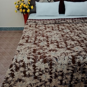 May include: A brown and cream floral patterned kantha quilt with a white background. The quilt is folded on a bed with white pillows and a brown headboard.