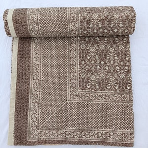 May include: A folded quilt featuring a brown and cream color palette with intricate block-printed patterns. The design includes a central section with floral motifs, surrounded by geometric borders and a solid cream-colored edge strip.