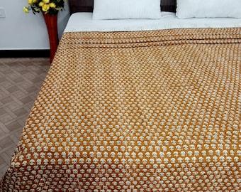 May include: A bed with a brown patterned quilt and two white pillows. The quilt has a repeating floral design in white on a brown background. A vase of yellow flowers sits to the side.