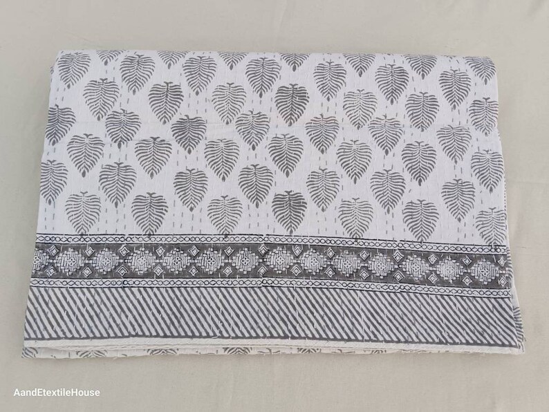 May include: A white cotton quilt with a grey leaf pattern and a grey and white border. The quilt is folded in half and lying on a white surface.