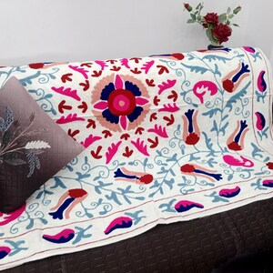 May include: A white embroidered sofa throw with a large central floral design in pink, red, and blue. The throw is draped over a brown sofa, with a decorative pillow featuring a floral design. A vase of red flowers sits in the background.