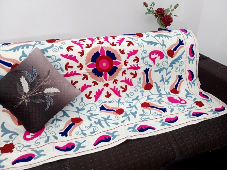 May include: A white embroidered sofa throw with a large central floral design in pink, red, and blue. The throw is draped over a brown sofa, with a decorative pillow featuring a floral design. A vase of red flowers sits in the background.