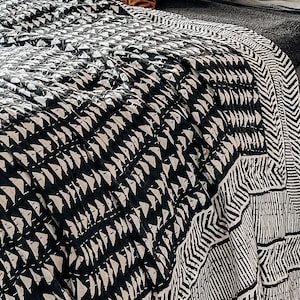 May include: A black and white patterned quilt with a geometric design is draped over a bed. The quilt features a repeating pattern of triangles and lines. A brown and two gray pillows are on the bed.