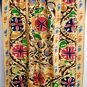 May include: A beige textile with intricate floral embroidery in shades of pink, blue, green, and brown. The design features large, stylized flowers and scrolling vines, creating a vibrant and detailed pattern. The edges are bordered with a repeating leaf and floral motif.