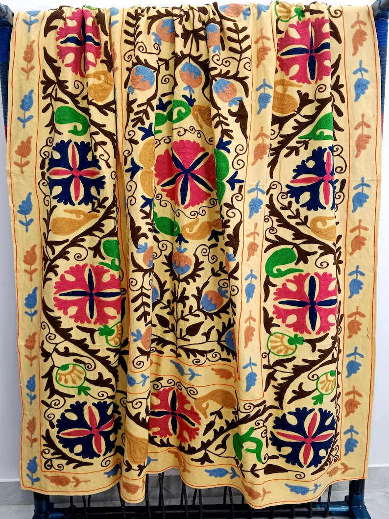 May include: A beige textile with intricate floral embroidery in shades of pink, blue, green, and brown. The design features large, stylized flowers and scrolling vines, creating a vibrant and detailed pattern. The edges are bordered with a repeating leaf and floral motif.