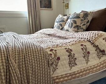 May include: A bed with a white bed skirt, a patterned quilt, and decorative pillows. The quilt features a floral design in burgundy and olive green on a white background. A textured, beige throw blanket is draped over the bed.